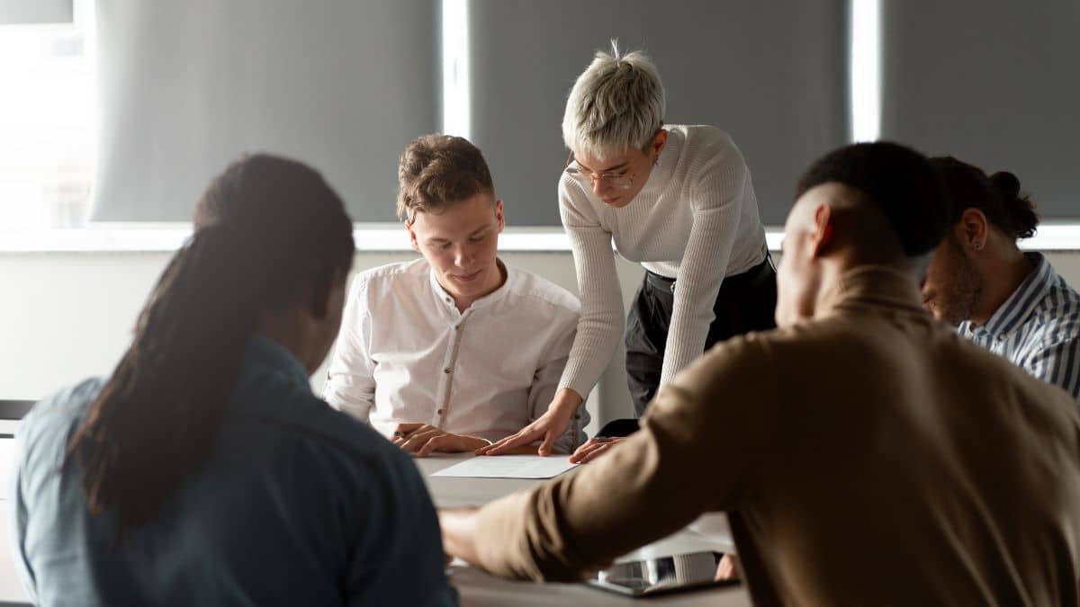 A group of five diverse individuals is engaged in a collaborative meeting. A person with short hair points at a document, fostering teamwork and focus.