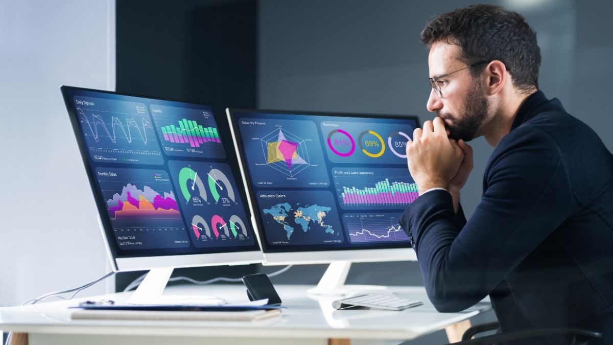 A Fractional CFO in a blue suit intently analyzes data on two computer monitors displaying colorful charts, graphs, and world maps in a modern office setting.